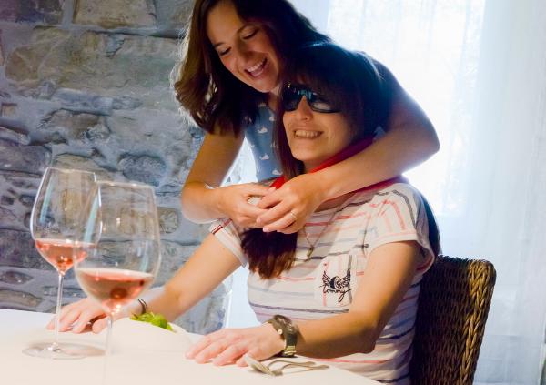 In a restaurant a girl ties a red scarf to another blind girl who is sitting at the table while they both laugh